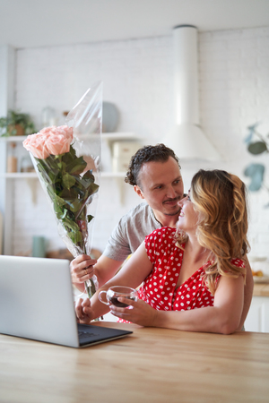 Surprise. Beautiful romantic couple in kitchen. Young man is presenting flowers to his beloved. Feel of happiness.の写真素材