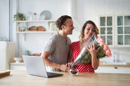 Surprise. Beautiful romantic couple in kitchen. Young man is presenting flowers to his beloved. Feel of happiness.の写真素材