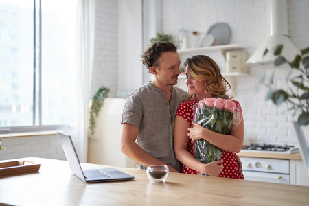 Surprise. Beautiful romantic couple in kitchen. Young man is presenting flowers to his beloved. Feel of happiness.の写真素材