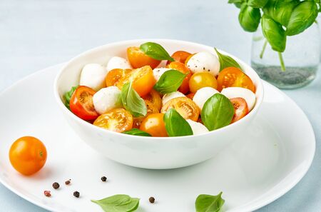 Caprese salad with yellow and red tomato cherry and baby mozzarella cheese in white bowl on white plate. Bocconcini mozzarella cheese with basil leaves.の写真素材