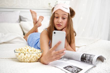 A young girl lies in bed after sleeping and browses social networks on the phone. Emotion is a surprise. Free time outside of school. Quarantine life.の写真素材