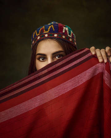 Art portrait of young beautiful girl with an embroidered hat on her head. The girl covers her face. Central Asian or oriental style, tradition and modernity. Beauty and fashion concept.の写真素材
