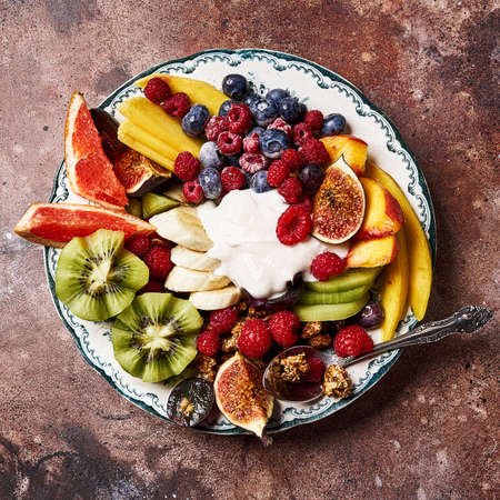 Assorted sliced fruits on vintage plate. Mango, kiwi fruits, figs, grape, bananas, raspberries and blueberries with yoghurt and granola.の写真素材