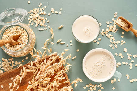 Oat milk in a glass and mug on a blue background. Flakes and ears for oatmeal and granola on a wooden plate.の写真素材