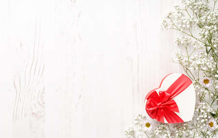 Flowers composition with gift and empty space for your text. White flowers on white wooden background and shaped heart gift. Wedding mockup with small flowers and gift with a red bow. Flat lay, top view, frame. Gypsophila Baby's-breath flowersの写真素材