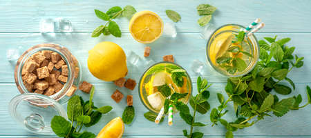 Summer cold drinks in glasses with lemons, mints on blue wooden background. Top view of lemonade...