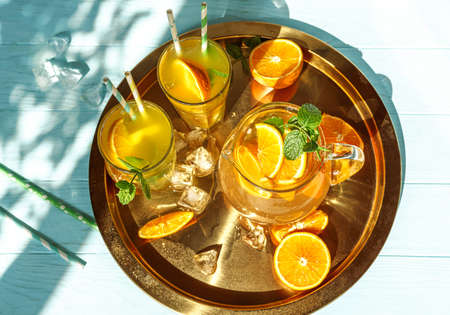 Glasses of lemonade with oranges, ice cubes and mint leaves on golden plate. Top view of lemonade on blue wooden background. Summer cold drinks under direct sun shines. Caustic reflection effect.の写真素材