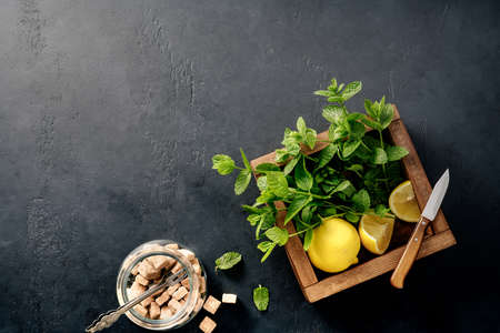 Fresh mint in wooden box on dark concrete background with lemons and cane sugar cubes. Top view of a...