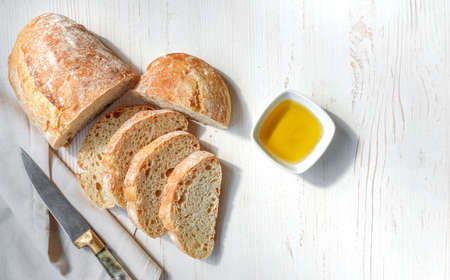 Sliced organic bread ciabatta with vintage linen napkin and bowl of olive oil on white wooden background. Top view of homemade ciabatta with copy space. Flat lay.の写真素材
