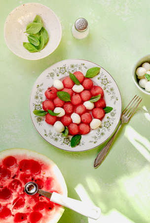 Watermelon salad with mozzarella cheese and fresh basil leaves on green background. Caprese salad with watermelon balls and spoon. Top view flat lay.の写真素材