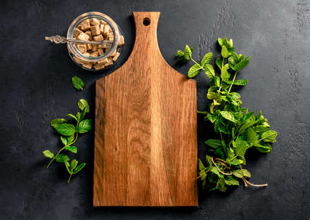 Rustic wooden board with fresh mint and cane sugar in glass bowl on dark black concrete background....