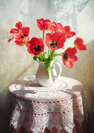 Rustic still life with flowers on a table covered with a white crocheted tablecloth. White jug with with red tulips on round table.の写真素材