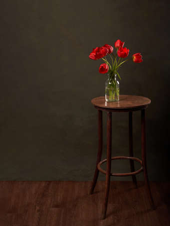 Vase with flowers on vintage table. Round table with red tulips on dark green background with copy space.の写真素材