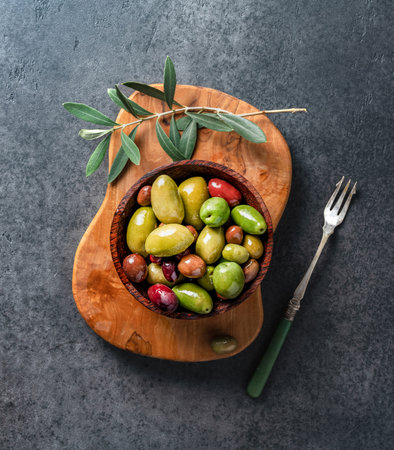Different olives in wooden bowl on olive board with olive branch. Tasty olives on dark background.の写真素材