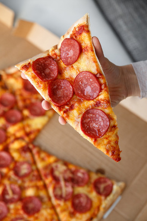 Hand holding a piece of pepperoni pizza. Hot homemade food. Sliced of fresh italian classic salami pizza. Popular topping with cheese. Baked meal.の写真素材