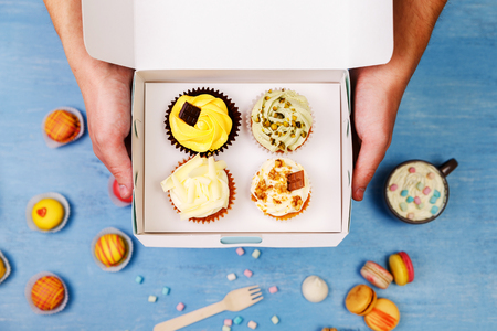 Cupcakes and macaroons. Male hands holding delivery box. Cakes with buttercream. Almond macaron cookies. Sweet dessert.の写真素材