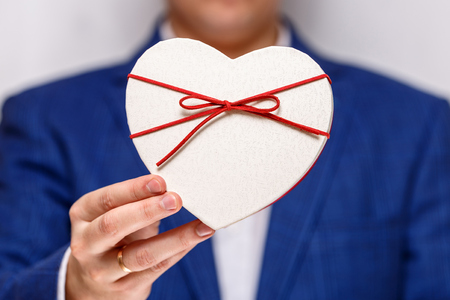 Male hands holding heart-shaped gift box. Present wrapped with ribbon and bow. Valentines day love package. Man in suit and white shirt.の写真素材