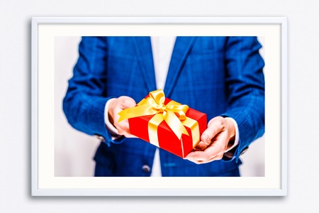 Male hands holding a gift box. Present wrapped with ribbon and bow. Christmas or birthday red package. Man in suit and white shirt. Wall frame poster with gift box photo. Mockup template.の写真素材