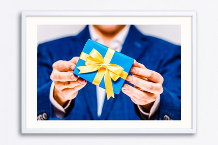 Male hands holding a gift box. Present wrapped with ribbon and bow. Christmas or birthday blue package. Man in suit and white shirt. Wall frame poster with gift box photo. Mockup template.の写真素材