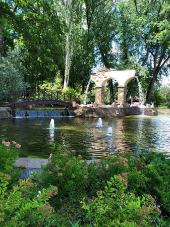 Landscape with a beautiful park in which there is a wooden bridge with an arch and water jets, with a pond, a fountain, trees and flowers on a sunny summer day.の写真素材