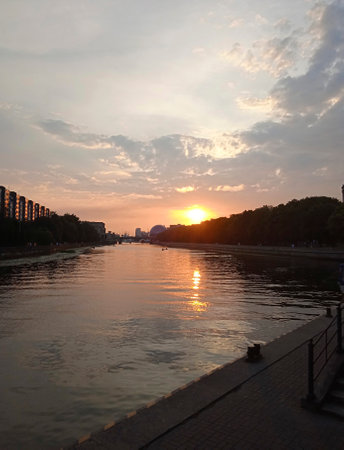 A beautiful sunset on the river in the city is visible from the embankment. On the left you can see houses in the distance. The houses are illuminated by the setting sun. The trees on the right in the distance are already in the twilight of the evening.の写真素材
