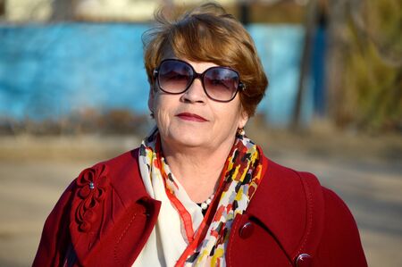 Portrait of stylish 67 year old woman in a bright red coat and sunglasses kindly and sincerely smiling on a spring dayの写真素材