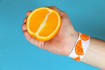 Male hand with woven handmade friendship bracelet holding half of ripe orange on blue backgroundの写真素材