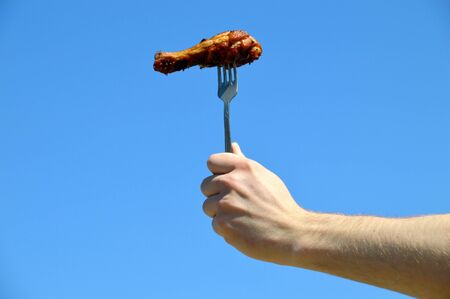 Hand holding fork with grilled appetizing chicken leg against the blue sky. Outdoor barbecueの写真素材