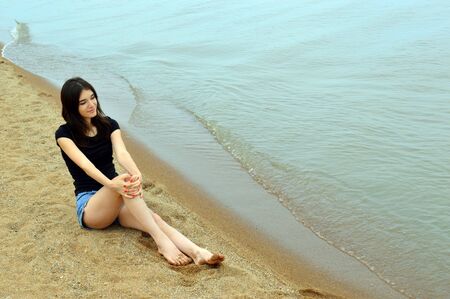 Attractive brunette girl with red nails sitting near water and looking away on a cloudy summer day. Lake Khanka, Russia.の写真素材