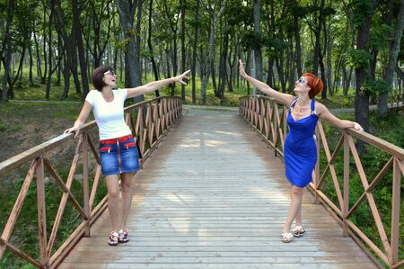 Two beautiful sisters stretching hands to each other and laughing sincerely on the wooden foot bridgeの写真素材