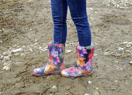 Women's legs in blue jeans and floral rubber boots on dirty soilの写真素材