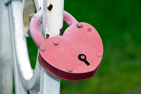 Red heart-shaped padlock hanging on a metal white fence on green background