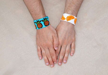 Males hands with woven friendship bracelets with alpha pattern pineapples and oranges fruit handmade of thread on beige backgroundの写真素材