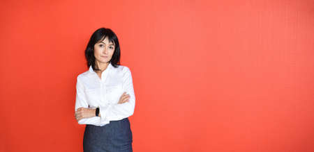 45 y.o. Russian brunette woman in white shirt with crossed arms looking at the camera and standing against red background. Success and business concept. Copy spaceの写真素材