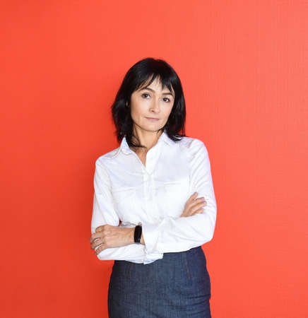 45 y.o. Russian brunette woman in white shirt with crossed arms looking at the camera and standing against red background. Success and business concept.の写真素材