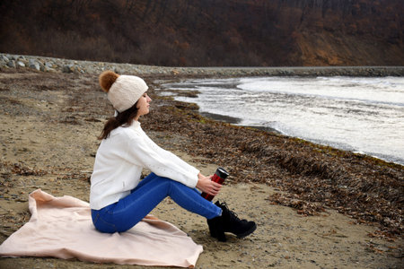 Young woman in white sweater, hat with pom-pom, sitting on the shore of the Japanese sea with closed...