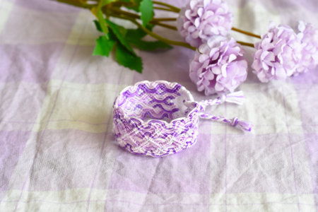Purple DIY friendship bracelet with unusual braiding on lilac textile next to flowersの写真素材