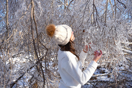 Portrait of young Russian woman in white sweater and beanie next to frozen branches of birch trees,...