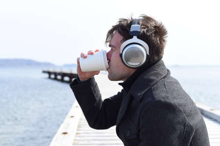 Young man listening to music and drinking coffee, sitting alone on pier on a sunny dayの写真素材