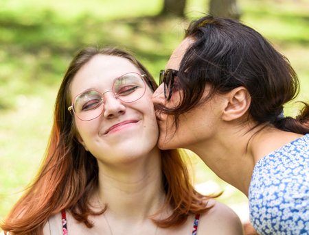 Mother kissing her teenage daughter with dyed hair and glasses against of green leavesの写真素材