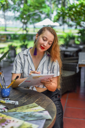 Beautiful blonde woman artist and illustrator thinking about a concept of her drawing, sitting by the table with drawings, brushes and paintの写真素材
