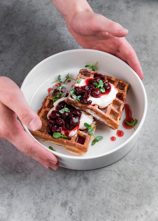 Waiter serving white plate with sweet belgian waffles with cream and cherry jam on a gray table in a cafeの写真素材