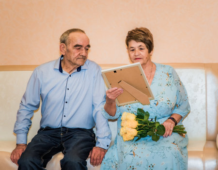 76-year-old man and 71-year-old woman celebrate their 50th wedding anniversary. The woman in a blue dress holds yellow roses, and they look at a gift a frame with a diploma and photoの写真素材
