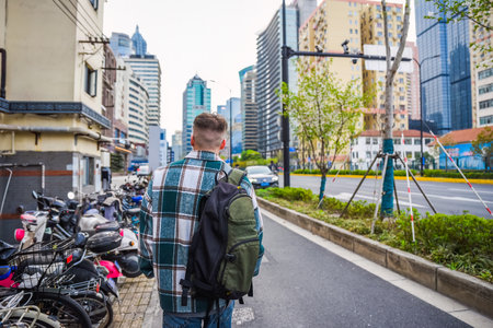 Russian male tourist with backpack explores modern Shanghai alone, walking past scooter, car, and high-rise building on city streetの写真素材