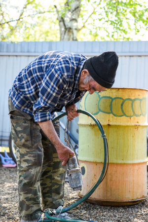 Elderly Russian man standing by a barrel with a dry pump in his hands, checking the hose before work in a quiet rural yardの写真素材