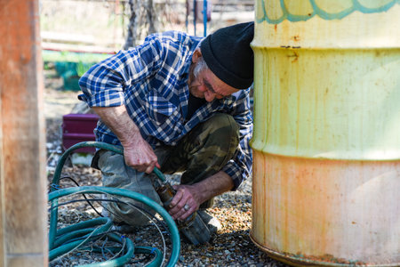 Elderly Russian man sitting near a barrel pulling a drainage hose to the side with visible tension and effort in a quiet rural setting.の写真素材