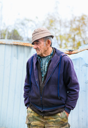 Elderly Russian man standing in a rural village, hands in pockets, looking sideways, dressed in...