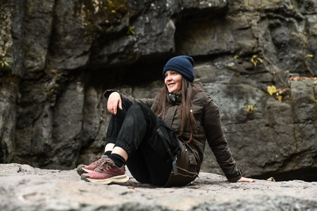 Woman traveler sitting on a rock near a waterfall. Autumn hike adventure in the mountains, enjoying nature and outdoor freedom.の写真素材
