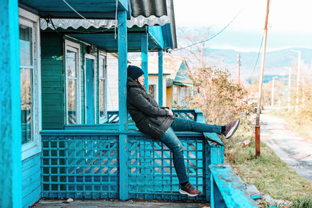 Woman sitting on the blue wooden porch of an old rural house in a village. Autumn day, calm...