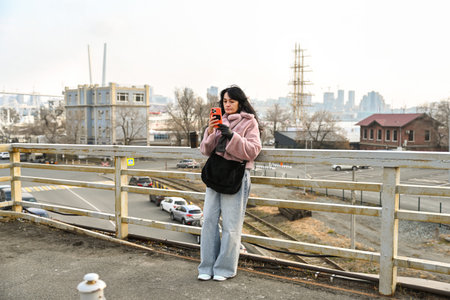 A 50 year old Russian woman stands on an overlook in Vladivostok using her smartphone, enjoying a calm urban morning with soft light and a city view.の写真素材
